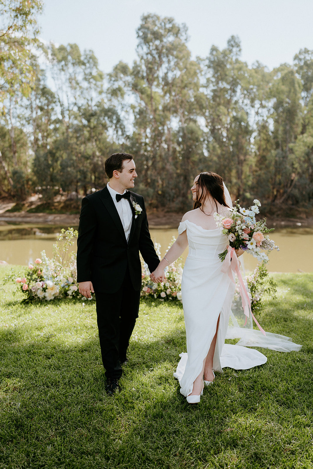 Wedding couple walking together with a scenic background of trees and water, holding a custom made wedding bouquet by Taylor Made Blooms.