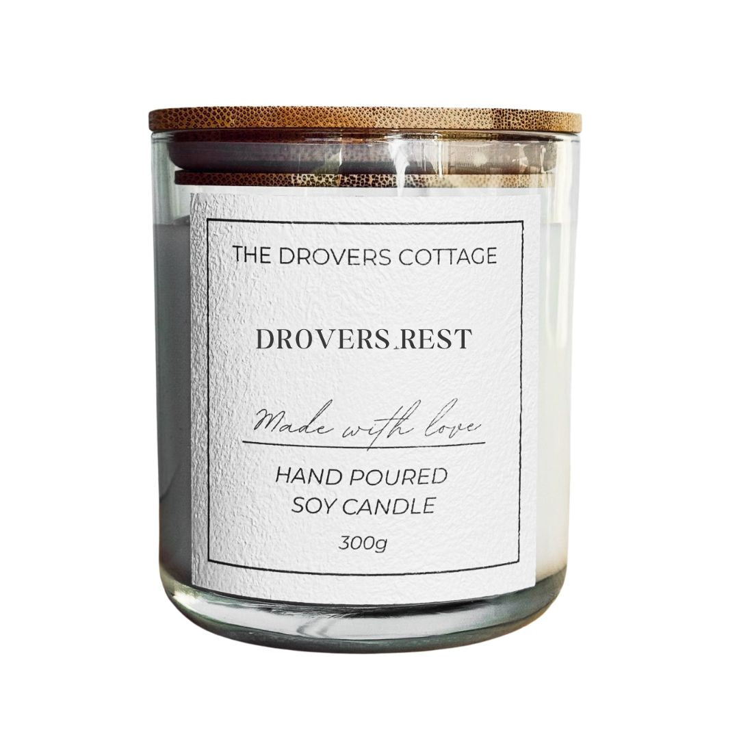 Candle jar with wooden lid and 'The Drovers Cottage' label on a white background