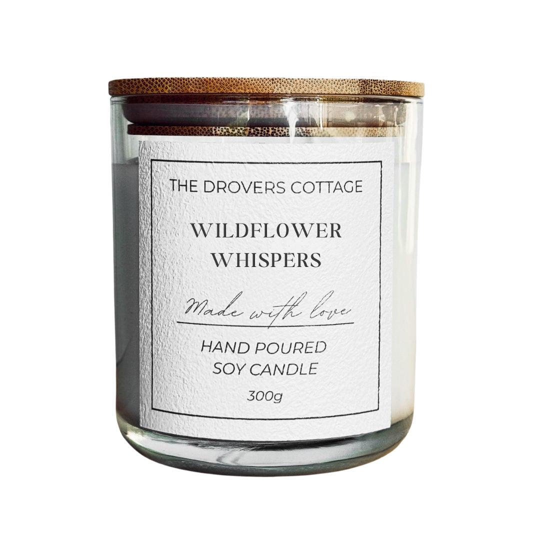 Candle with a wooden lid labeled 'The Drovers Cottage Wildflower Whispers' on a white background