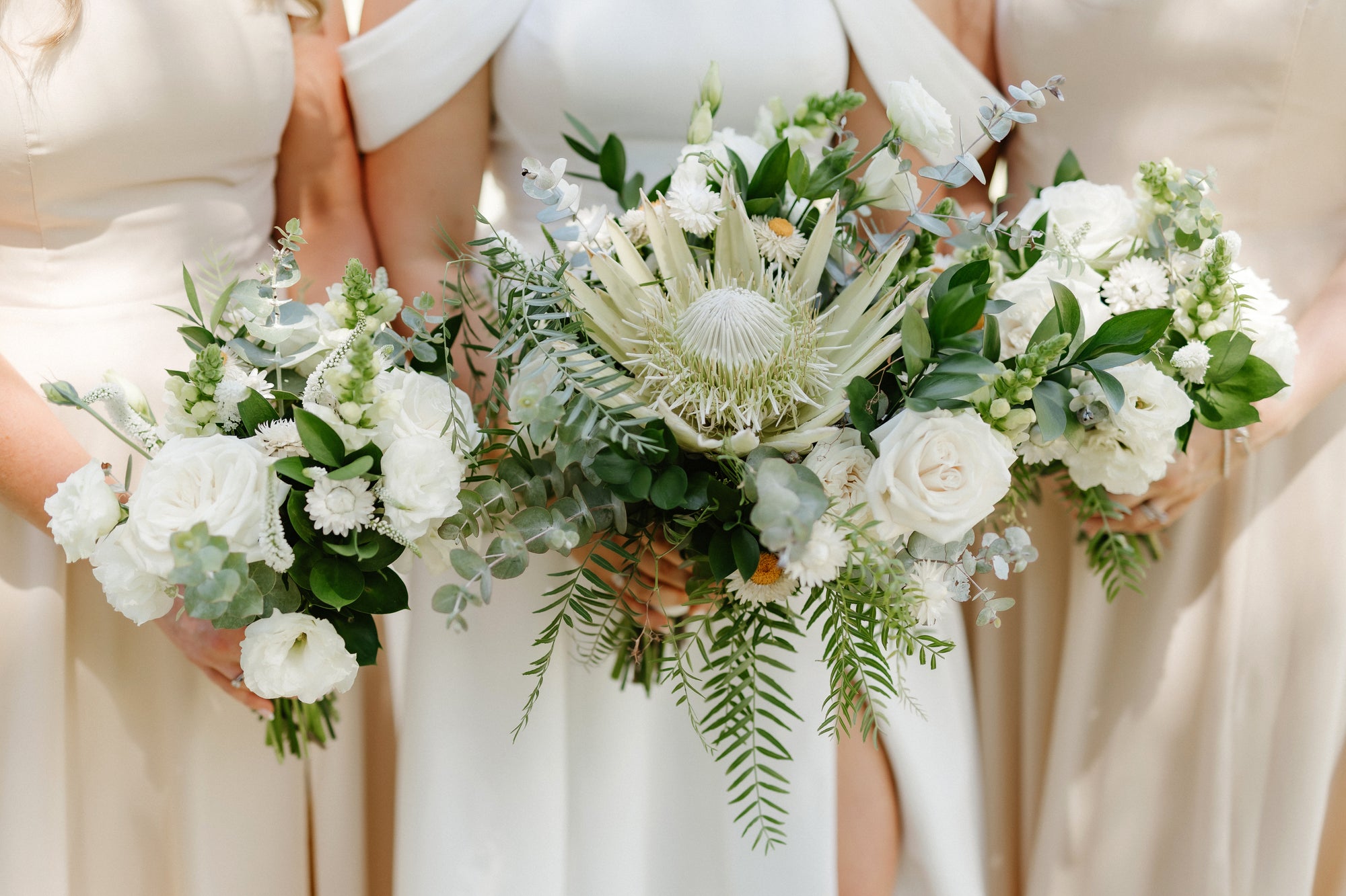 Three women holding bouquets of white and green flowers, wearing white dresses.